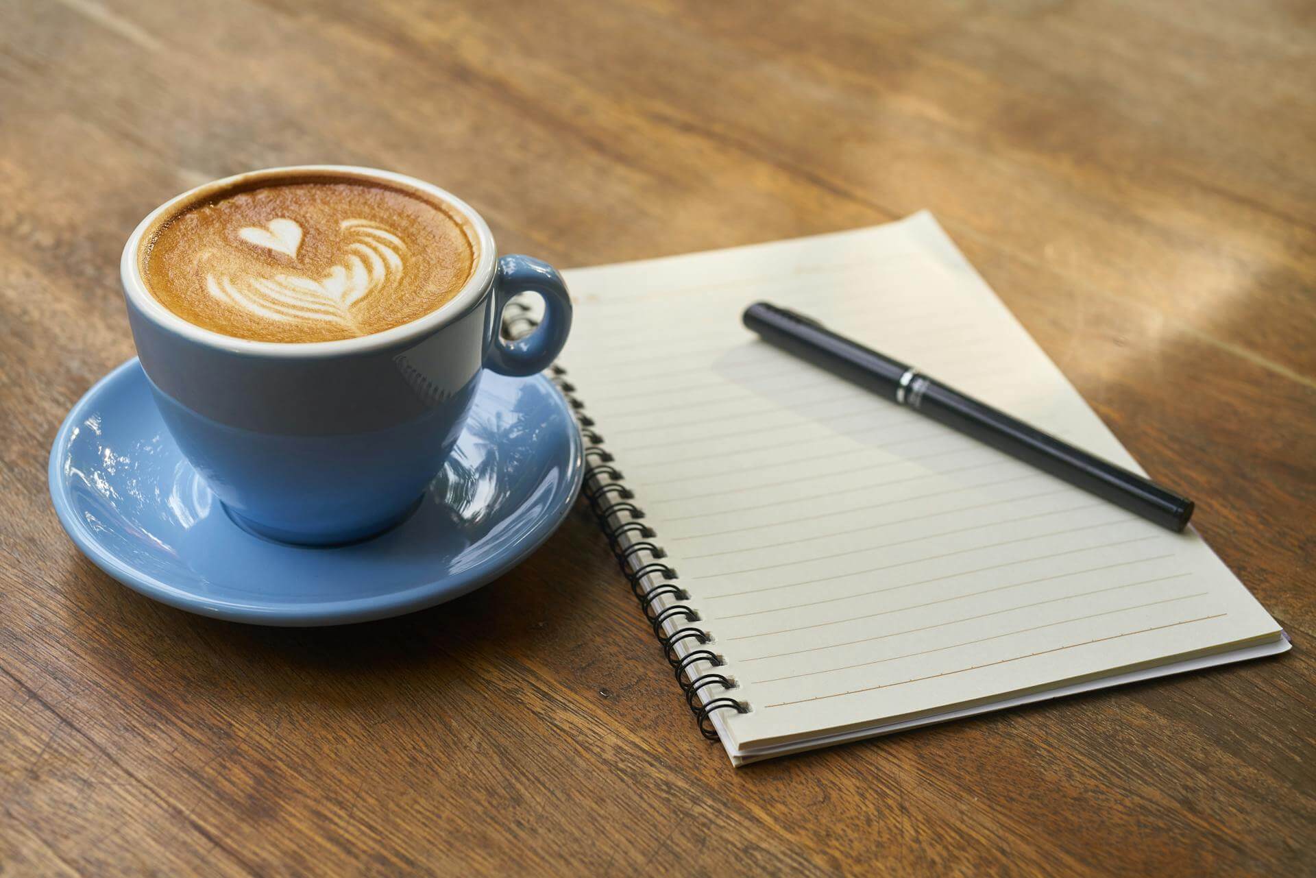 Coffee in a blue cup and saucer beside a notebook and pen