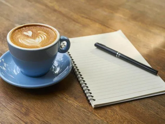 Coffee in a blue cup and saucer beside a notebook and pen