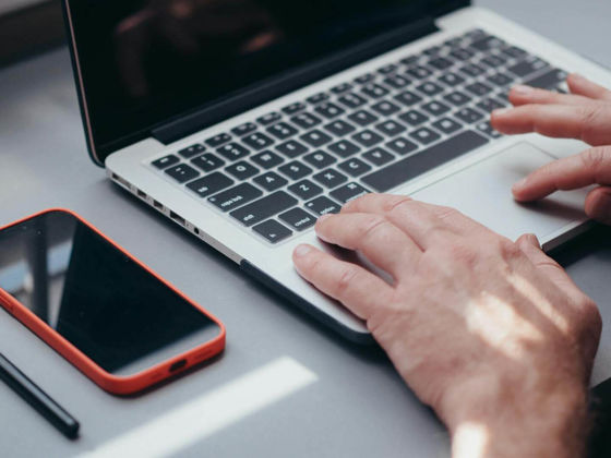 A person using a laptop with a smart phone on the desk next to them (Pexels - kindelmedia)