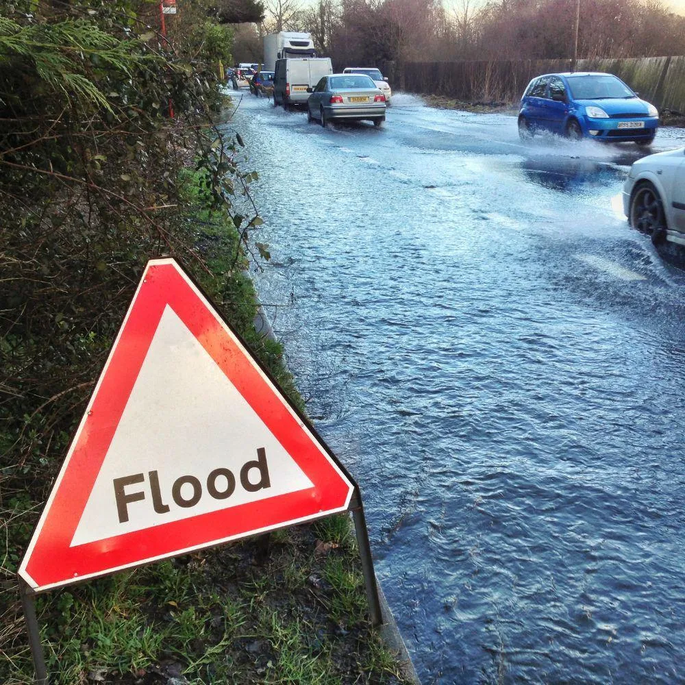 Flood warning sign beside a partially flooded road with cars driving through water and trees lining the roadside