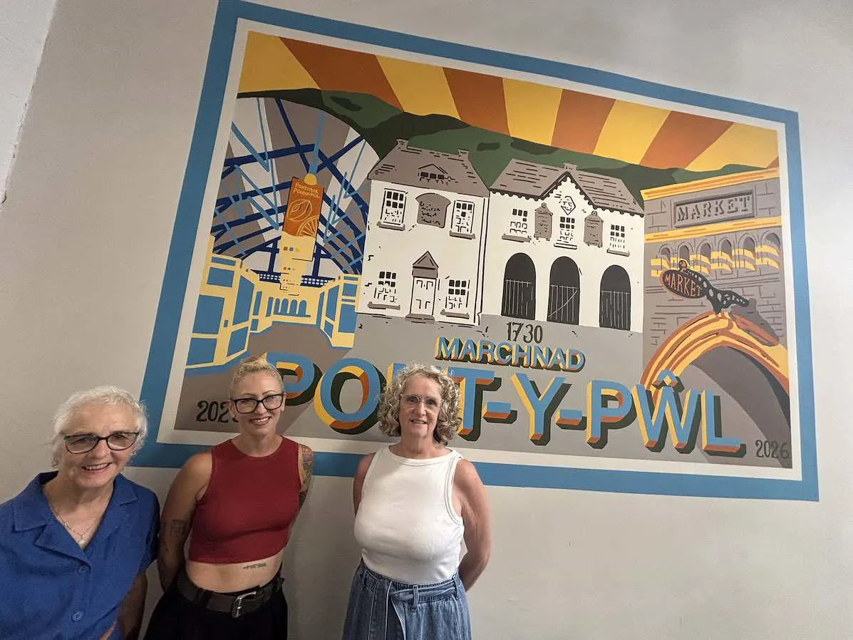 Three people standing in front of a colourful Pontypool Market mural featuring local landmarks, market architecture and the Welsh text "Marchnad Pont-y-pŵl", with sunrise-style artwork in the background.