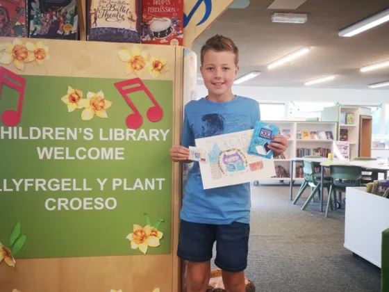 A young boy holding up his library card competition design and his new library card at Cwmbran Library.